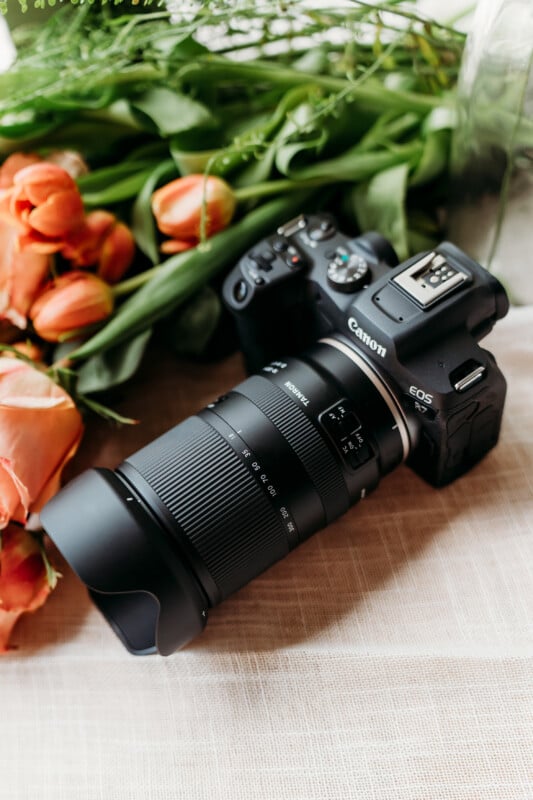 A Canon EOS R5 camera with a large zoom lens rests on a light fabric surface next to a bouquet of orange tulips and green leaves.