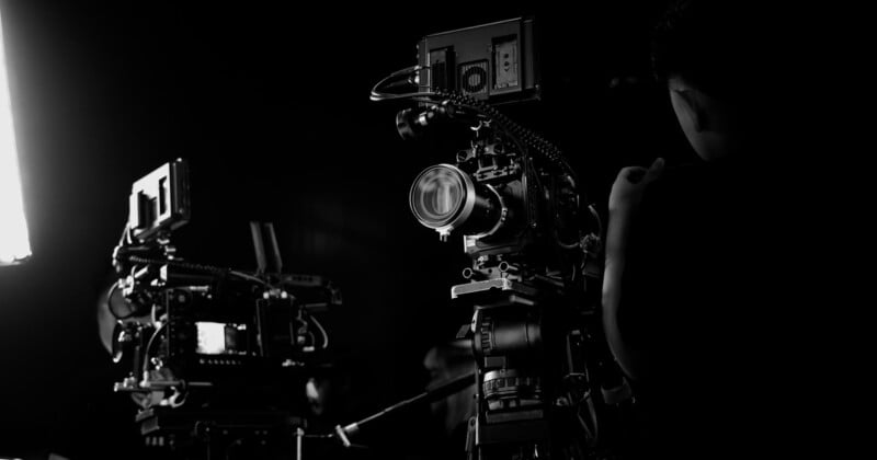 Una fotografía en blanco y negro de dos cámaras profesionales instaladas en un estudio oscuro y una persona parada junto a una cámara, con el lado derecho parcialmente visible. La luz brillante proviene del lado izquierdo de la imagen.