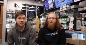 Two men sit inside an electronics store, both wearing hoodies with "impulse" logos. Shelves with gadgets, boxes, and brightly lit screens are visible in the background. One man has glasses and long red hair.