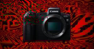 A Canon EOS R mirrorless camera is displayed against a bold red and black abstract patterned background. The camera’s lens mount is exposed, and the Canon logo is visible on top.