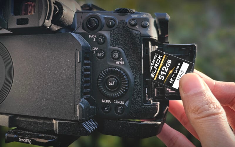 A hand inserts a 512GB V90 SD memory card into the open card slot of a professional black digital camera mounted on a tripod.