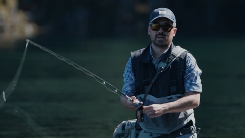 A man wearing sunglasses, a cap, and a fishing vest stands in shallow water while fly fishing, holding a fishing rod. The background is blurred, showing a natural outdoor setting.