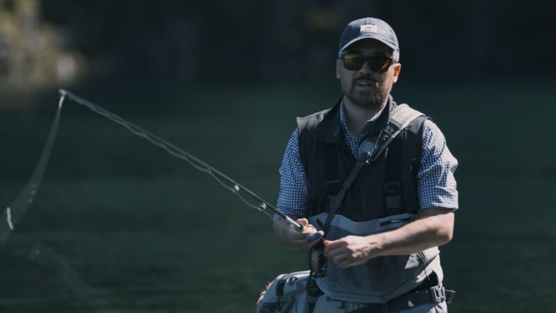 A man wearing sunglasses, a cap, and a fishing vest is fly fishing in a river or lake, holding a fishing rod with a cast line visible in the air.