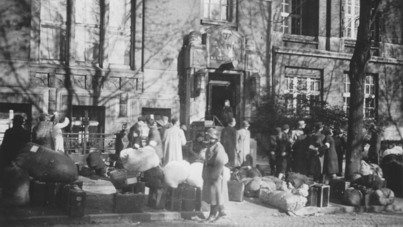 Una fotografía en blanco y negro muestra a personas reunidas afuera del edificio con maletas y paquetes; algunos hacen cola en la entrada, otros esperan en la acera, rodeados de sus pertenencias.