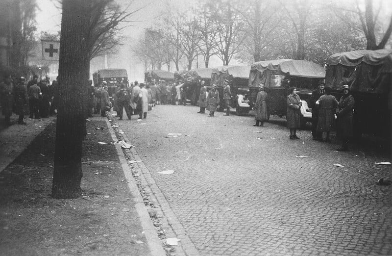Fotografía en blanco y negro de una calle adoquinada llena de camiones militares y soldados. La gente se reúne al fondo y se puede ver una cruz roja en el árbol de la izquierda. Las calles están bordeadas de árboles.