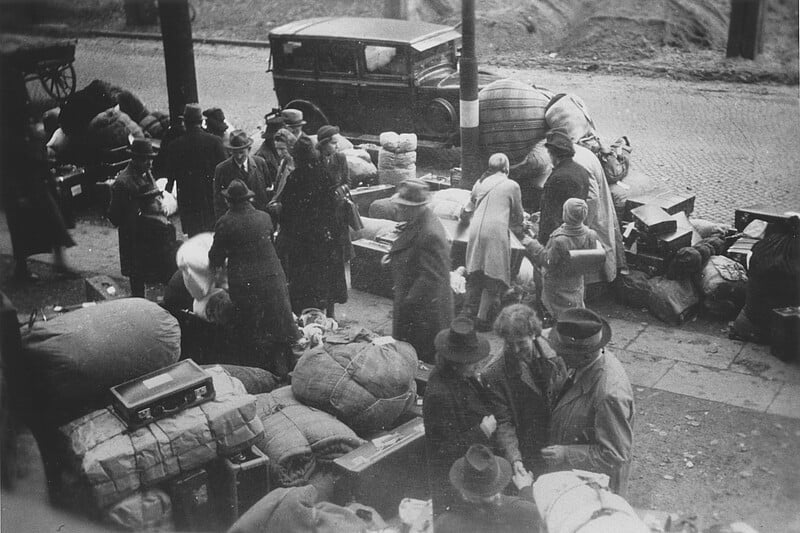 Un grupo de personas con sombreros y abrigos se reunieron alrededor de pilas de maletas y bolsos en la acera cerca de los autos estacionados, indicando que se estaban preparando o esperando en la carretera. La escena parece muy histórica.