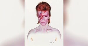 A person with pale skin and bright red hair styled upwards, eyes closed, with a bold pink and blue lightning bolt painted across their face. The background is plain white.