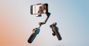 A smartphone mounted on a gimbal stabilizer with two detachable handles, filming a person indoors, against a gradient blue and orange background.