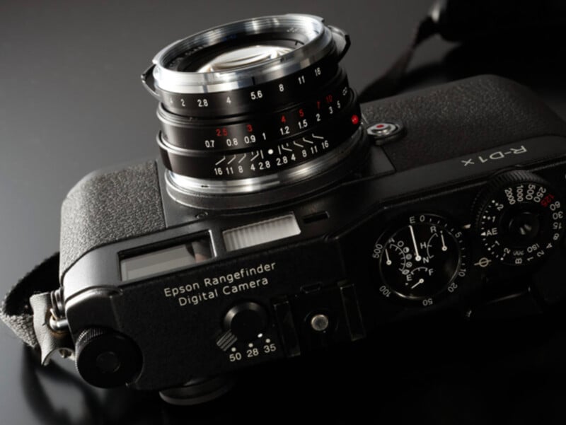 Primer plano de una cámara digital Epson Rangefinder negra con su dial, marcas de lentes y controles visibles sobre un fondo oscuro reflectante.