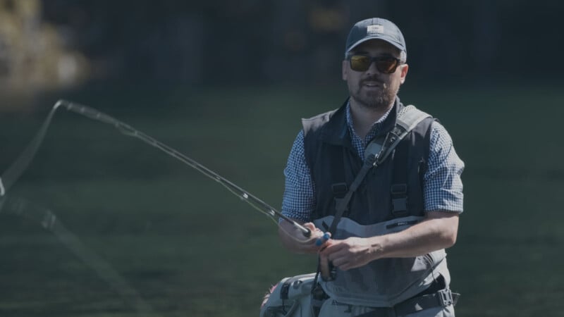 A man wearing sunglasses, a cap, and a fishing vest stands in a body of water, holding a fishing rod and casting his line, with a blurred natural background behind him.