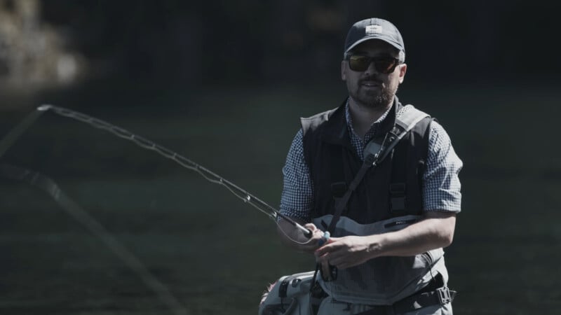 A man wearing sunglasses, a cap, and a fishing vest stands in water while holding a fishing rod, appearing to fish in a calm outdoor setting.