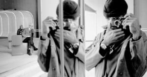 A person in a coat and beanie takes a photo with a camera in front of a mirror, creating a reflection. In the background, an older couple sits on a bench near a modern, curved structure. The image is black and white.