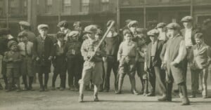 A group of boys in vintage clothing gather behind a chain-link fence, watching one boy stand ready to bat in a game of baseball on a dirt field, likely in the early 20th century.