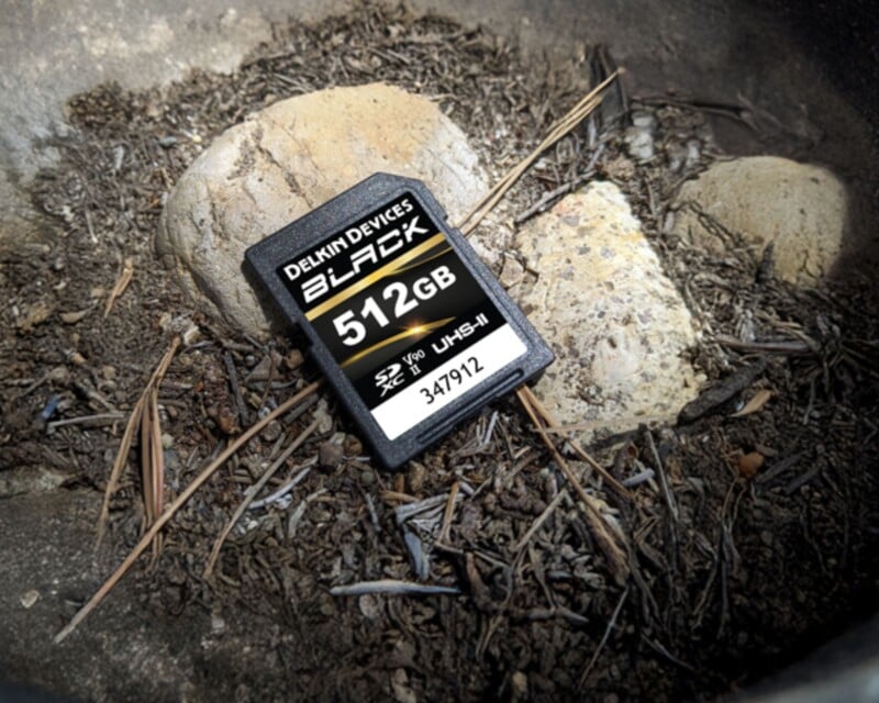 A 512GB Delkin Devices BLACK SD memory card lies on dirt and dried pine needles next to two rocks outdoors.