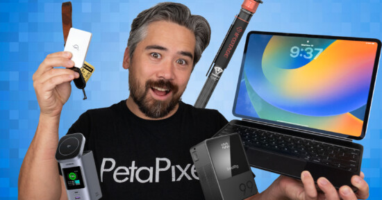 A smiling man in a "PetaPixel" shirt holds various tech gadgets, including an external drive, a camera, a battery, a tripod, and an Apple iPad with a keyboard, against a blue geometric background.