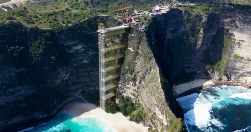 A tall glass elevator structure is built into a steep cliff, connecting a beach with turquoise water below to a small town above, surrounded by lush greenery and dramatic rocky cliffs.