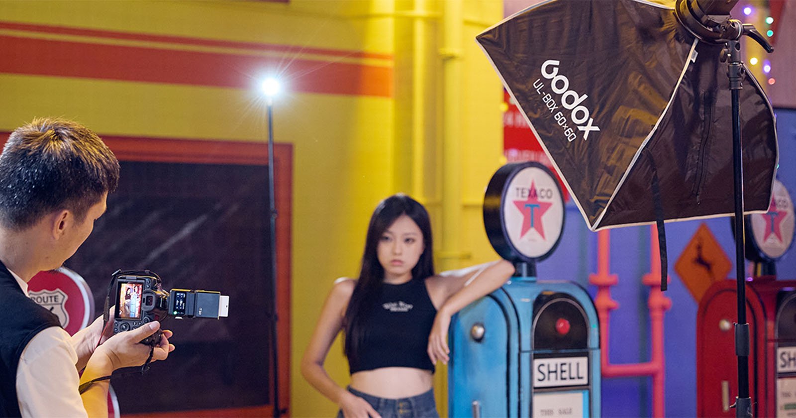 A photographer takes a picture of a woman posing in front of vintage gas pumps, with a large Godox softbox light set up nearby in a brightly colored indoor setting.