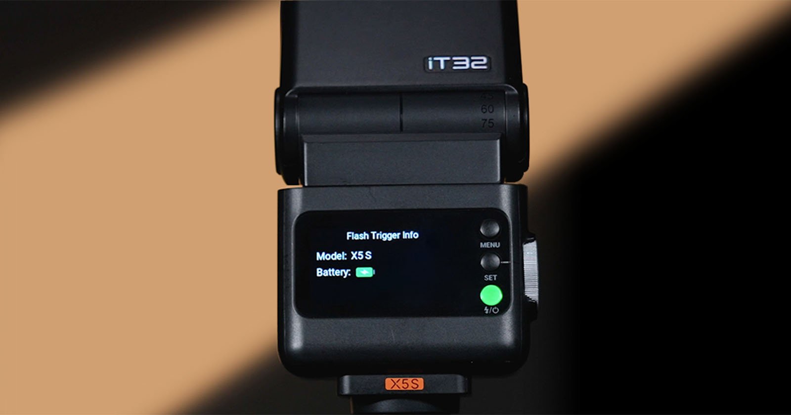 Close-up of a camera flash unit displaying "Flash Trigger Info" on its screen, showing mode as "XSS" and a full battery icon. The flash unit is black with buttons and a green LED light. The background is blurred beige.