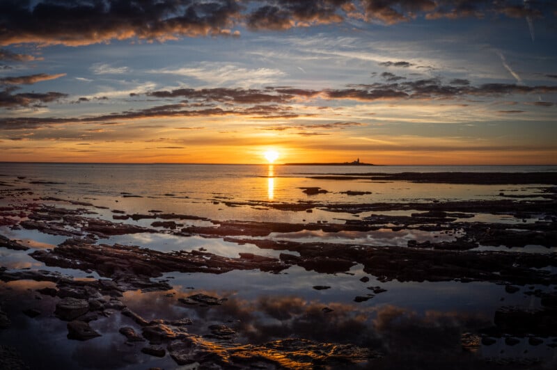 Colorida puesta de sol sobre el mar con nubes dispersas en el cielo y reflejos en la piscina en la costa rocosa. En el horizonte se ve claramente una isla con un faro, recortada contra la luz anaranjada del sol.