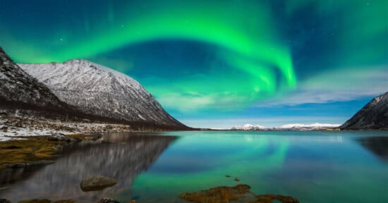 Green northern lights illuminate the night sky above snow-capped mountains and a calm lake, reflecting the vibrant aurora and surrounding landscape.