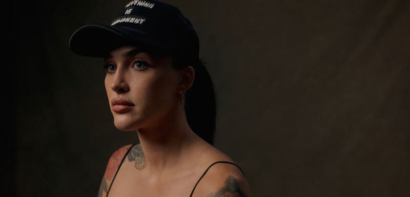 Una mujer tatuada con un sombrero negro y una blusa con tirantes finos se sienta sobre un fondo oscuro, mirando ligeramente de reojo con una expresión neutra.