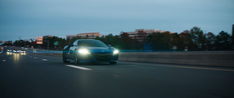 Un elegante auto deportivo negro con los faros encendidos acelera por una carretera casi desierta al anochecer, con árboles y edificios borrosos en el fondo.