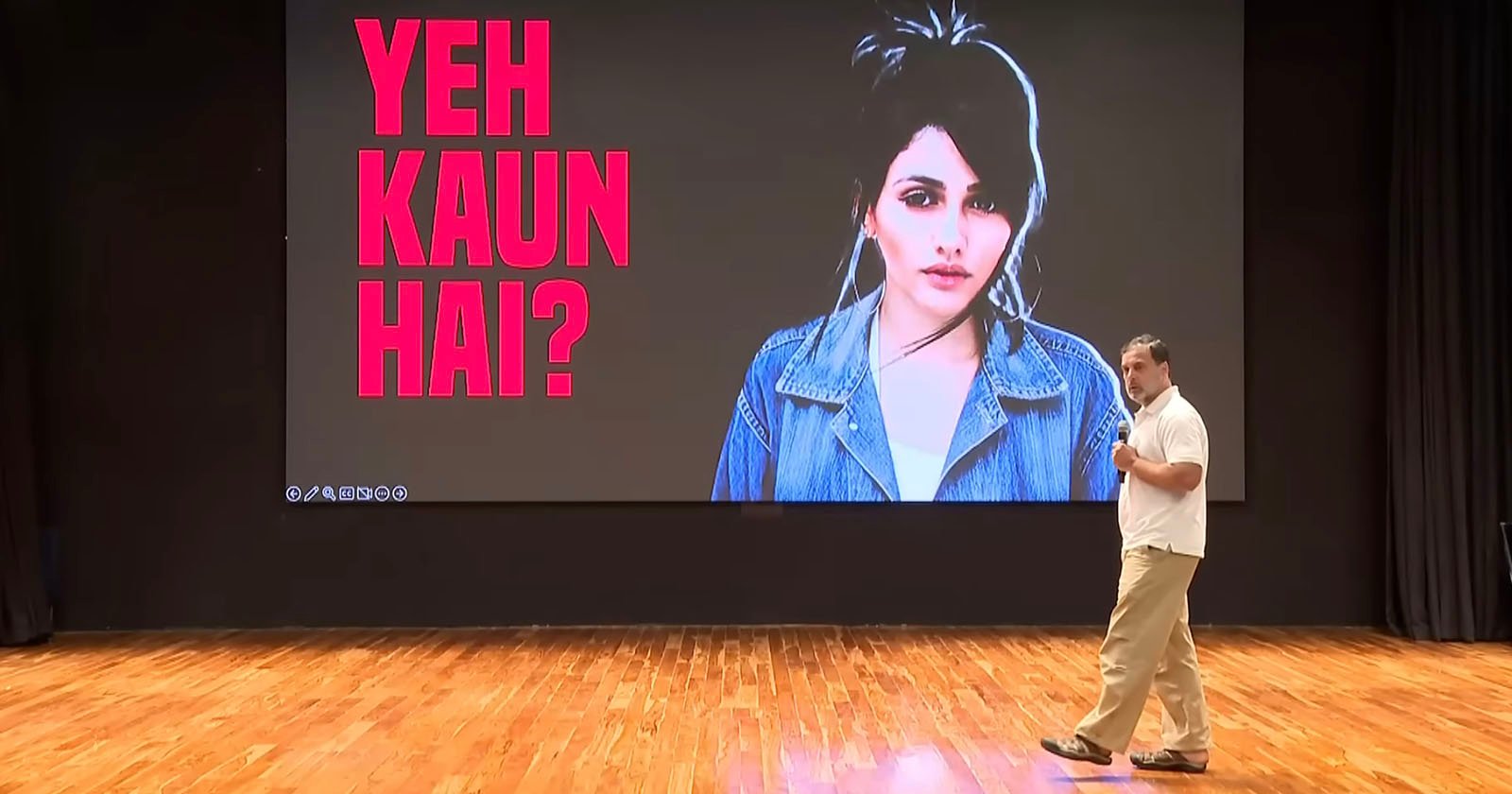 A person stands on a wooden stage in front of a large screen showing a woman’s image and bold pink text in Hindi reading "YEH KAUN HAI?" which means "Who is this?.