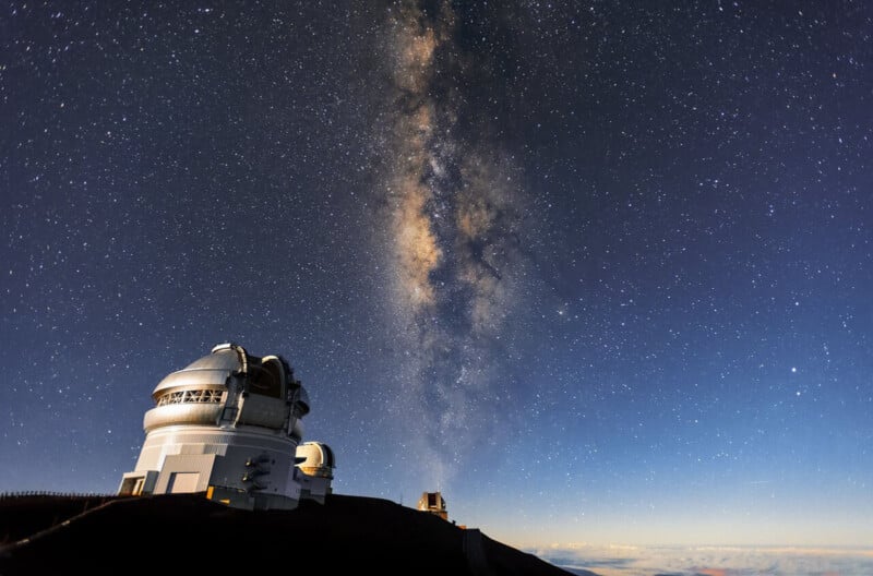 A night sky filled with stars and the Milky Way rises above a mountain-top observatory with domed telescopes, under clear conditions.