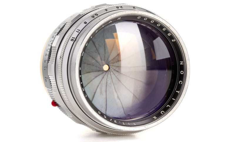 A close-up photo of a vintage Leica Noctilux camera lens, showing detailed aperture blades and engraved markings on the metal lens barrel, set against a white background.