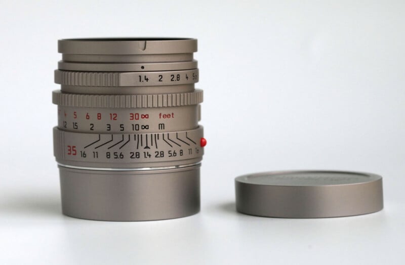A silver camera lens with engraved focus and aperture markings sits upright on a white surface next to its matching lens cap placed face down.