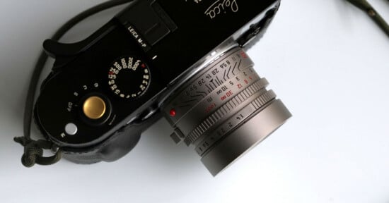 Close-up of a black Leica camera with a silver lens, showing detailed dials and engraved settings on both the camera body and the lens, placed on a white background.