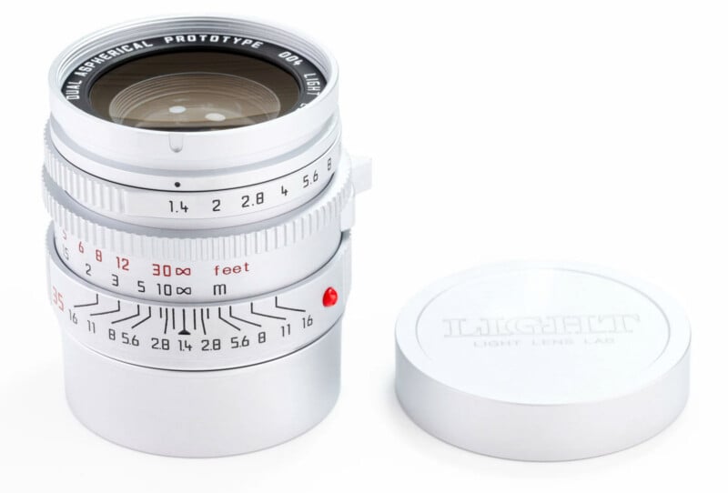 A silver camera lens with engraved markings sits upright beside a matching silver lens cap that reads “LIGHT LENS LAB.” The lens displays aperture and focus scales, with “DUAL ASPHERICAL PROTOTYPE” engraved on the front.