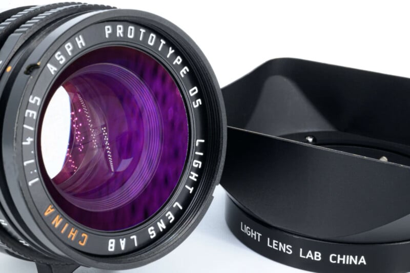 Close-up of a camera lens marked “ASPH PROTOTYPE 05 LIGHT LENS LAB CHINA 1:1.4/35” next to a detached square lens hood labeled “LIGHT LENS LAB CHINA.” The lens glass has a purple tint.
