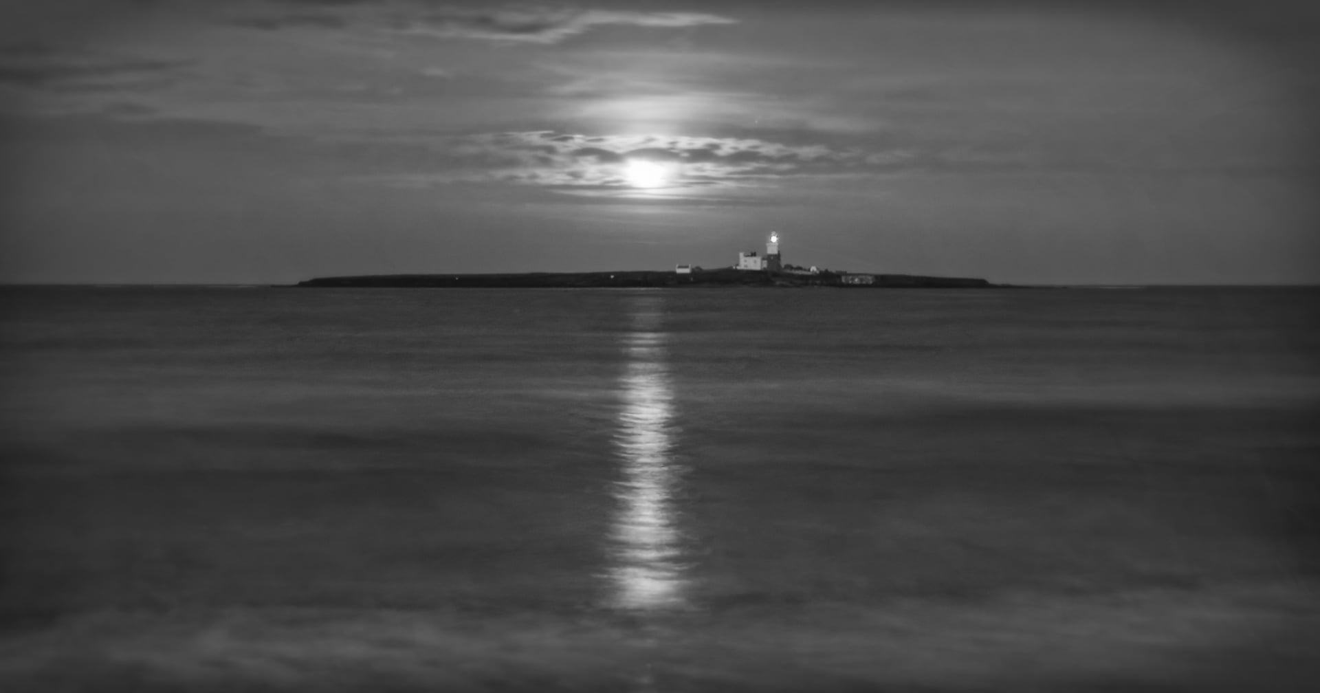 Una fotografía en blanco y negro de una pequeña isla con un faro, iluminada por la luz de la luna. La luna está parcialmente cubierta por nubes y su reflejo brilla en las tranquilas aguas del primer plano.