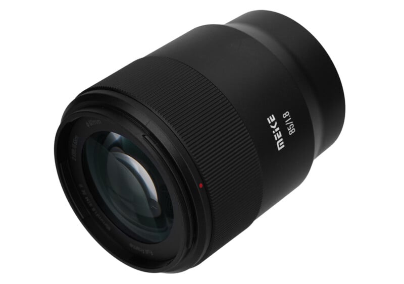 Una lente de cámara Meike 85 mm f/1.8 negra se muestra en ángulo sobre un fondo blanco, mostrando sus elementos de vidrio, su anillo de enfoque ranurado y la marca impresa en el cilindro de la lente.