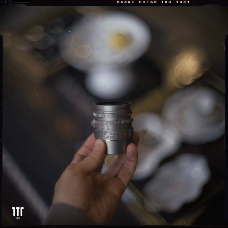 A hand holds a silver camera lens in focus, while the background features blurred white dishes and bowls with a soft, dreamy bokeh effect. The image has a vintage film aesthetic.