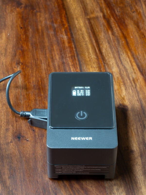 A Neewer battery charger sits on a wooden surface with a power cable plugged in. Its display screen shows battery level information in white text and icons.