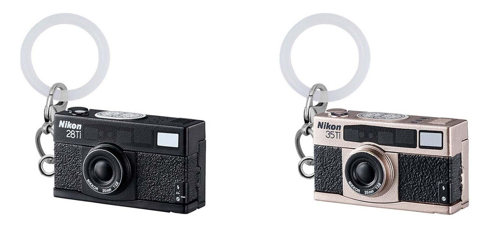 Two miniature camera keychains resembling Nikon 28Ti (black) and Nikon 35Ti (champagne silver) film cameras, each with a circular transparent keyring attached.