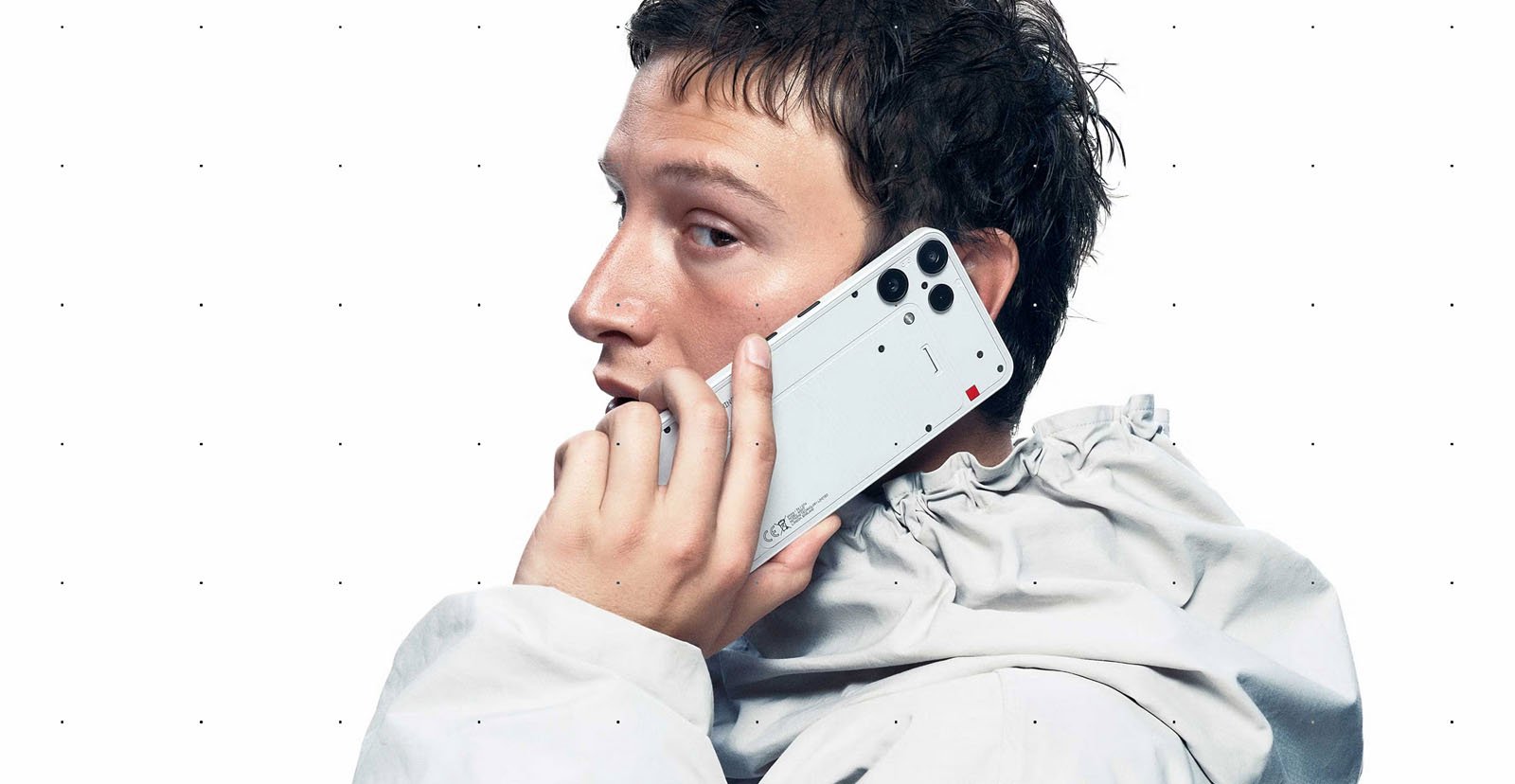 A person wearing a light gray, high-collared jacket holds a white smartphone to their ear, looking over their shoulder against a plain white background.