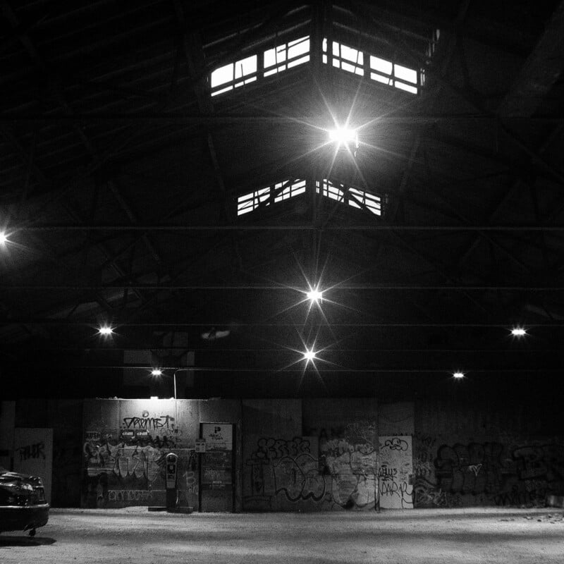 Una fotografía en blanco y negro de un almacén vacío y con poca luz, con paredes cubiertas de graffiti, luces brillantes en el techo y ventanas cerca del techo. A la izquierda se ve parte de un coche aparcado.