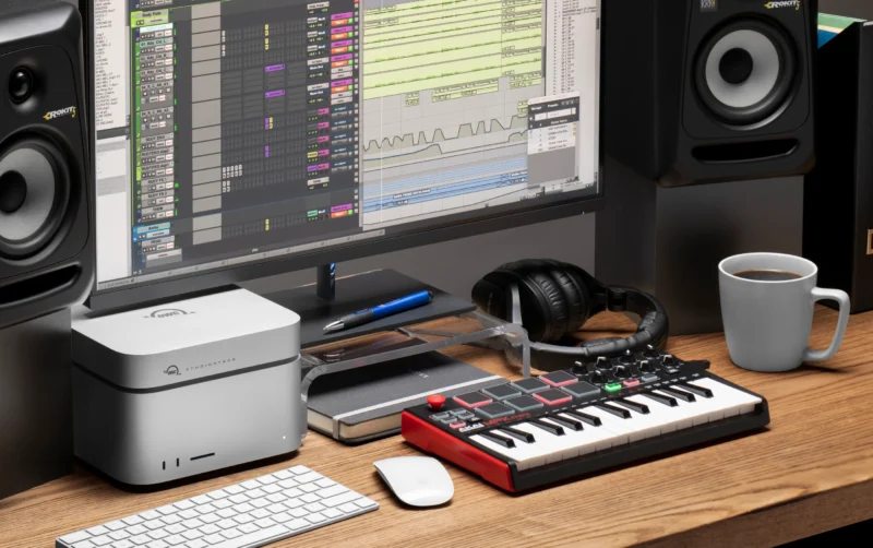 A modern music production workspace with a computer displaying audio editing software, studio monitors, a mini MIDI keyboard, headphones, a coffee mug, a notebook, and a pen on a wooden desk.
