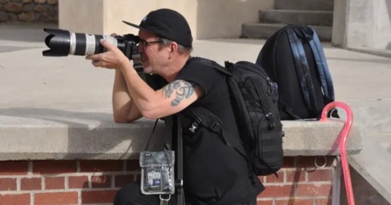 Un hombre con gafas, sombrero y mochila se arrodilla al aire libre y toma fotografías con una lente de cámara grande. Cerca hay una mochila negra y un bastón rosa.