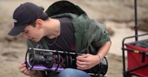 A person wearing a black cap and green jacket examines a cylindrical underwater camera housing while sitting outdoors on a sandy surface.