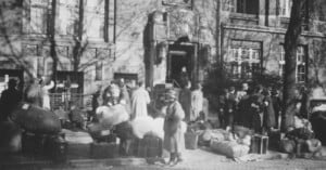 A black-and-white photo showing a group of people standing outside a building with piles of luggage and bags on the sidewalk; some individuals look towards the entrance while others wait or stand nearby.