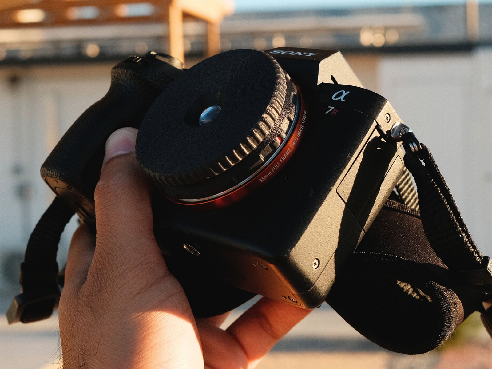 A close-up of a hand holding a Sony Alpha 7R camera, shown without a lens attached. The background is blurred, suggesting an outdoor setting with sunlight.