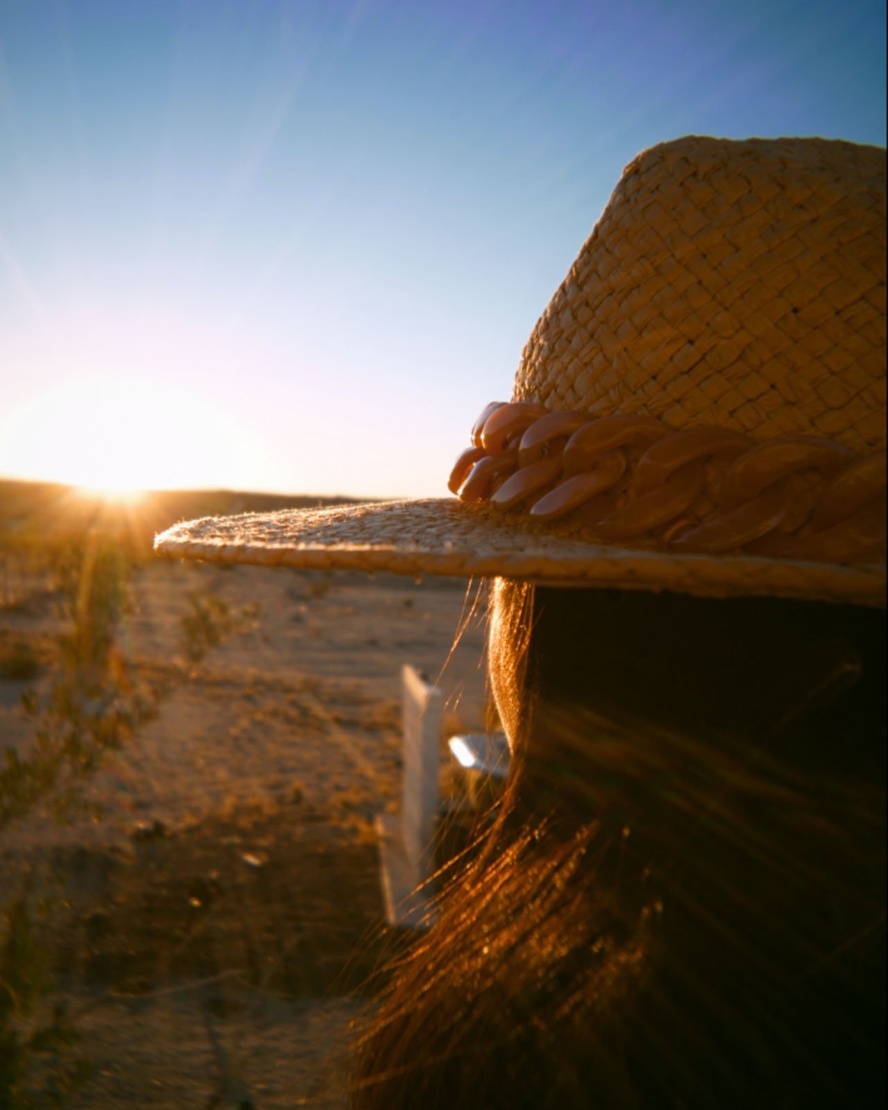 A person wearing a woven straw hat faces the setting sun in a desert landscape, casting a warm golden glow and long shadows across the sand.