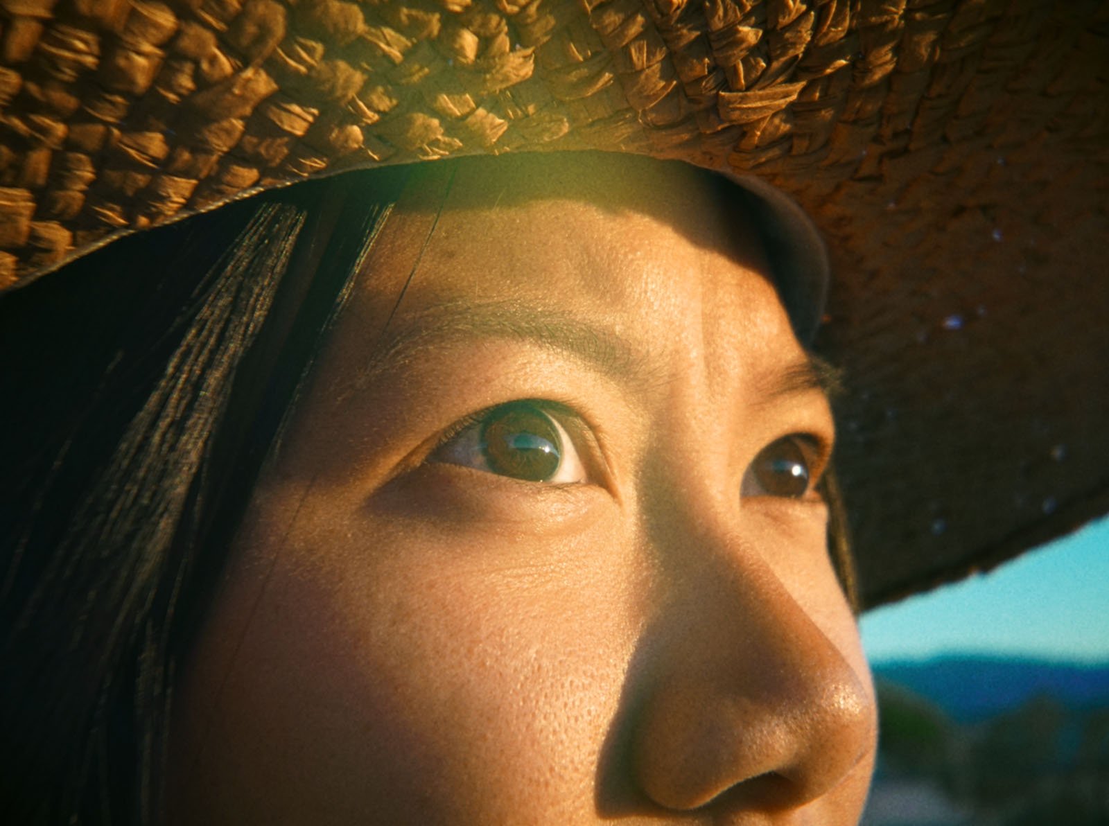 Close-up of a person's face in sunlight, focusing on their eyes and upper nose. The person is wearing a woven straw hat, and a warm, golden light enhances their skin. The background is blurred.