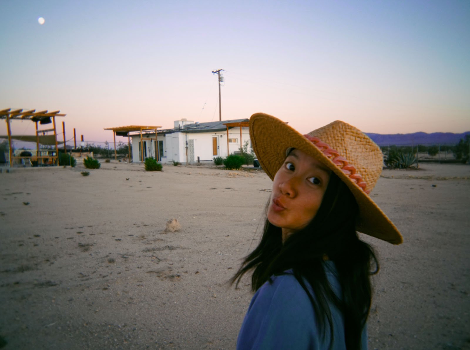 A woman wearing a wide-brimmed straw hat and a blue shirt looks back at the camera, making a playful face, standing in a desert landscape with sparse buildings and mountains in the background at sunset.