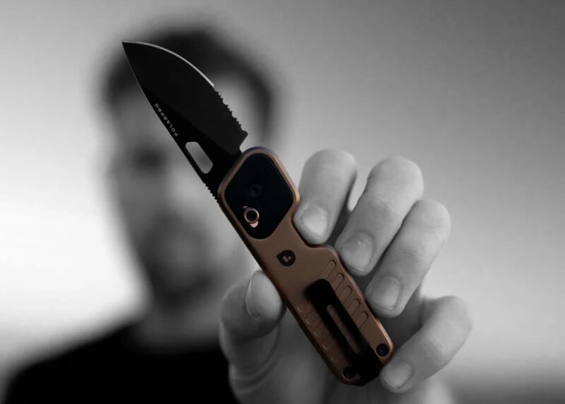 Un hombre sostiene una navaja plegable compacta con hoja negra y mango de color cobre y la sostiene hacia la cámara. El fondo está difuminado en escala de grises, destacando el cuchillo.