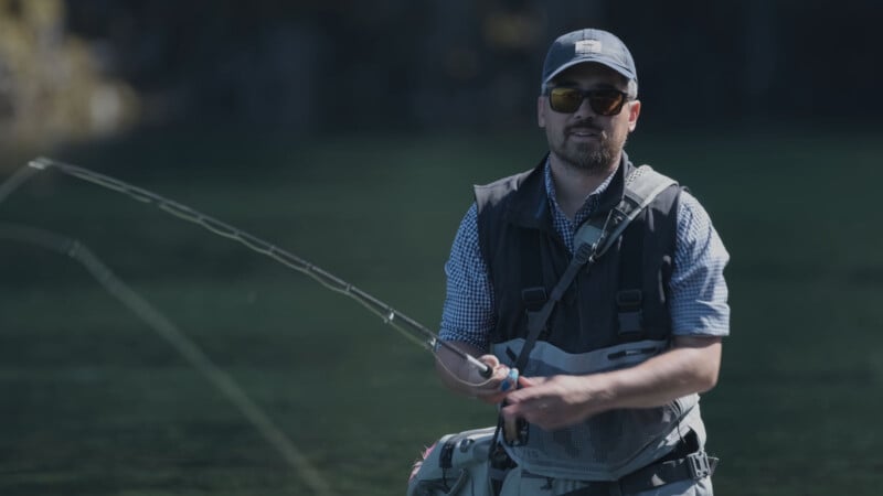 A man wearing sunglasses, a cap, and a fishing vest stands in shallow water, holding a fishing rod, with greenery and water blurred in the background.